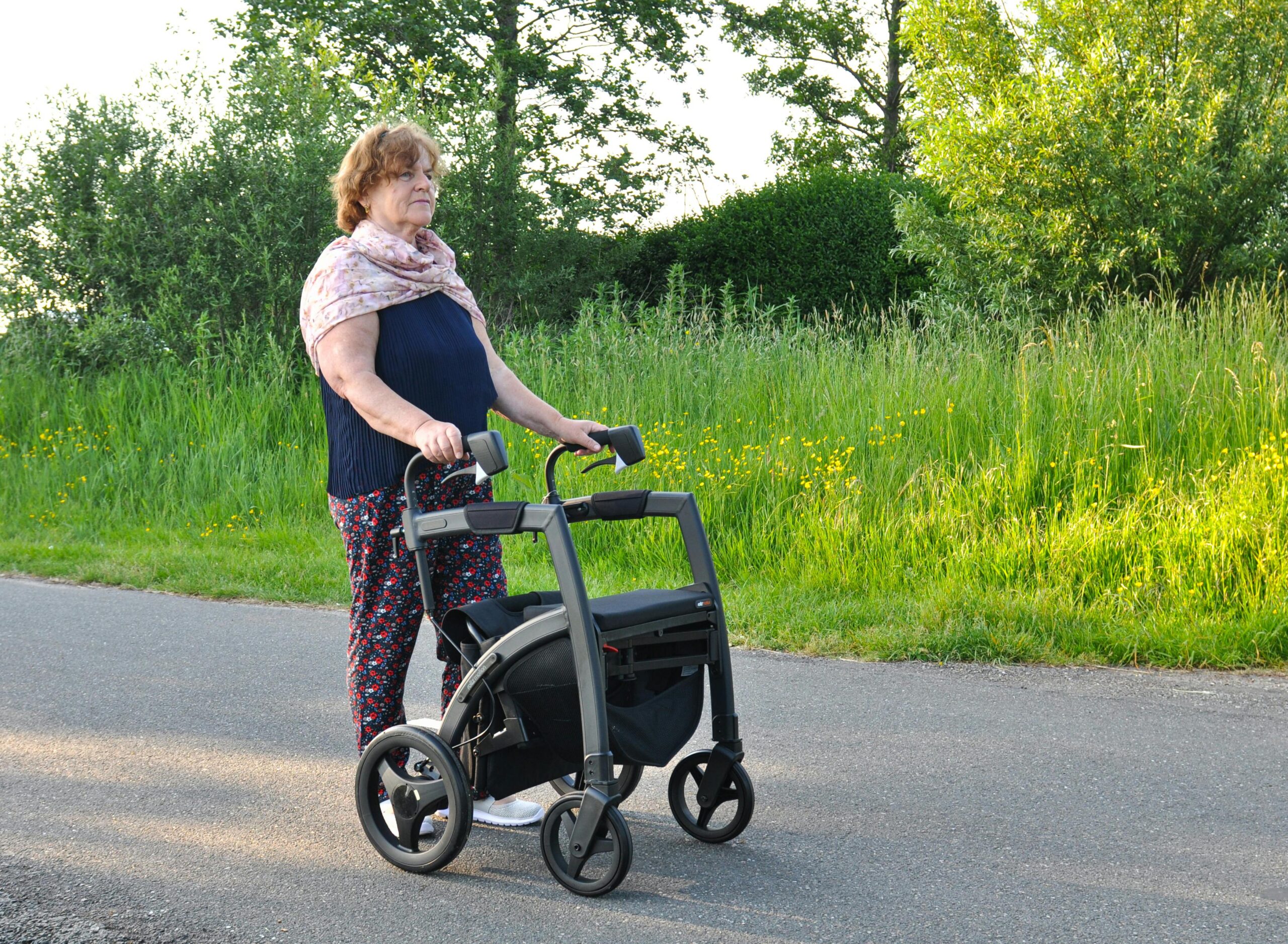 Senior woman enjoying a sunny day outdoors with a rollator walker, surrounded by green nature.