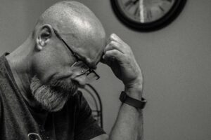 Black and white portrait of a thoughtful bald man indoors, capturing a moment of reflection with a wall clock in the background.