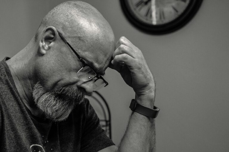 Black and white portrait of a thoughtful bald man indoors, capturing a moment of reflection with a wall clock in the background.