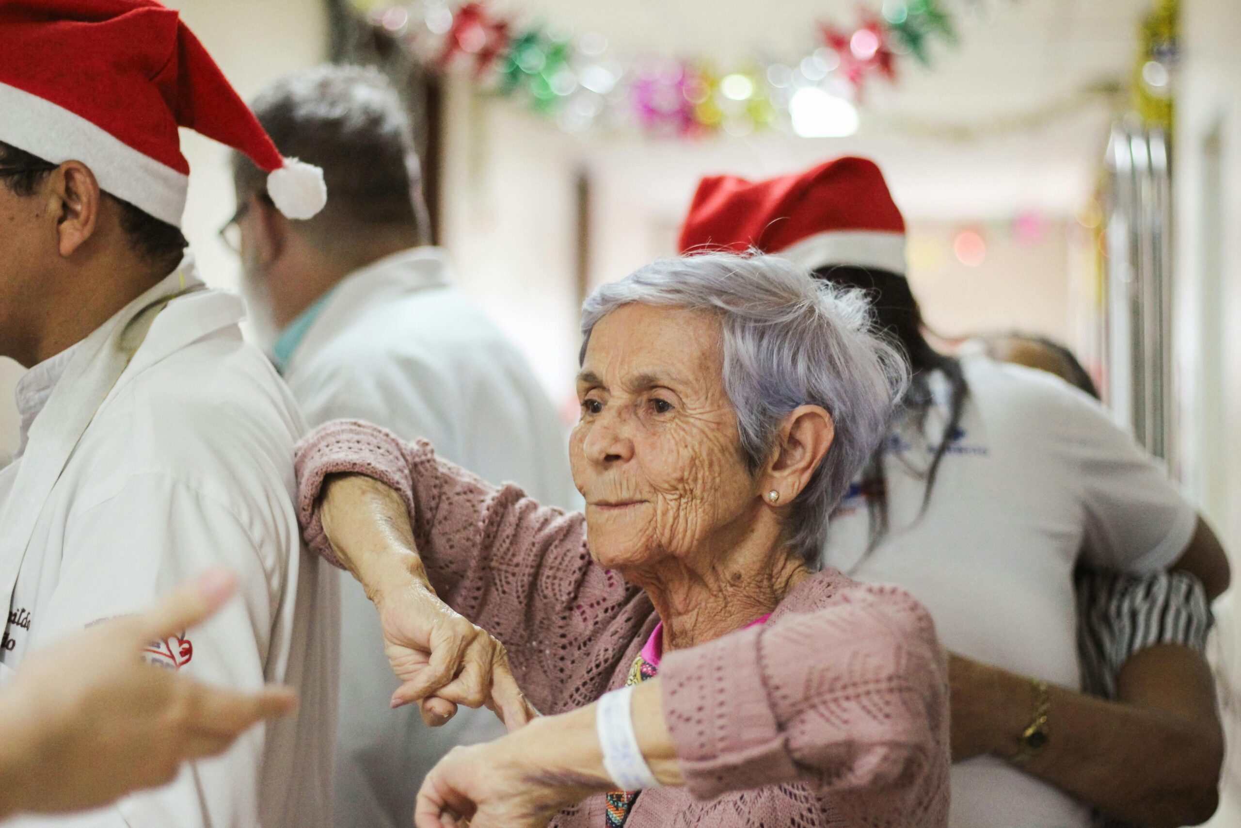 A joyful elderly woman in a festive setting, surrounded by people wearing Santa hats.